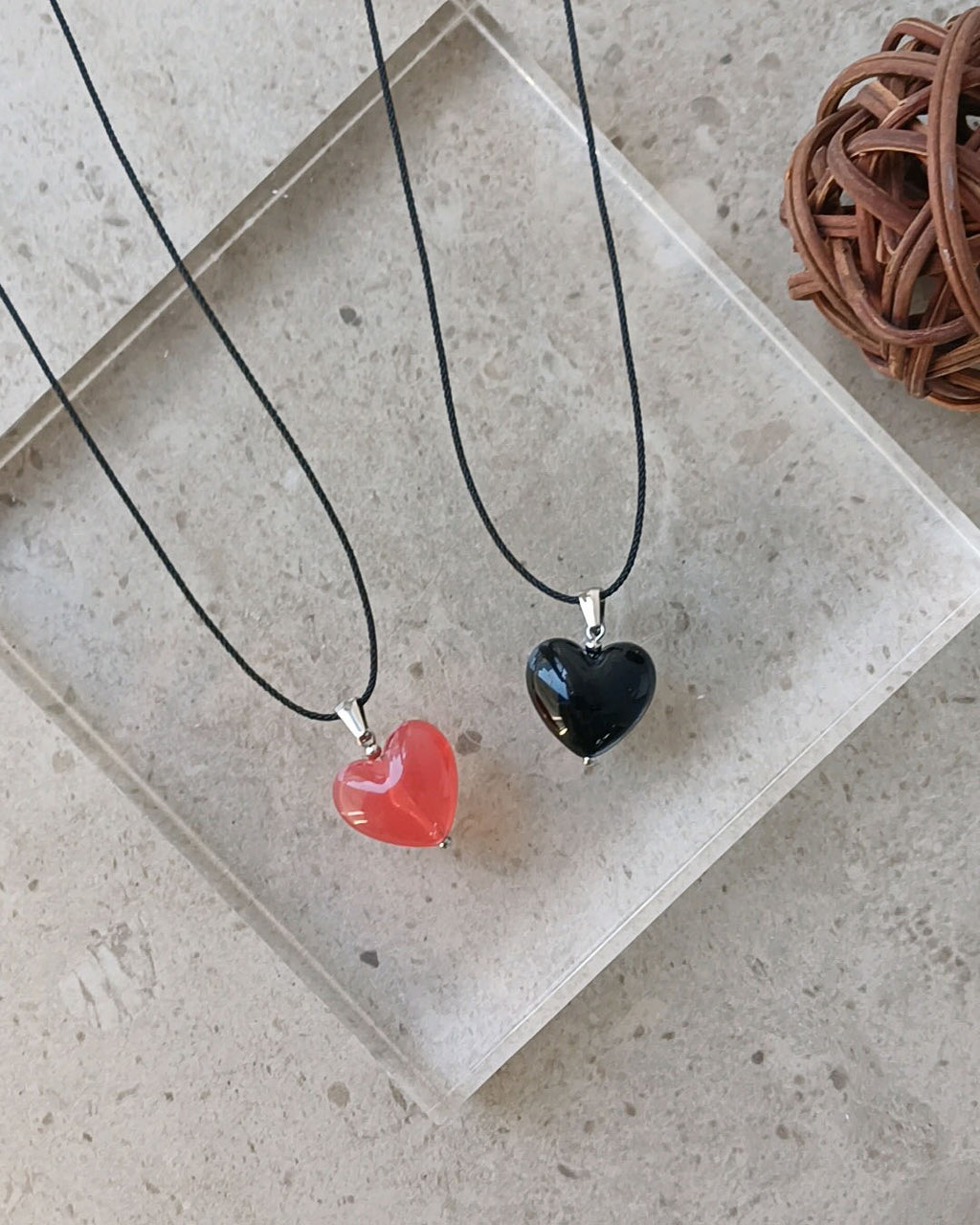 Two heart-shaped pendants on black cords with a concrete surface and wicker ball in the background.