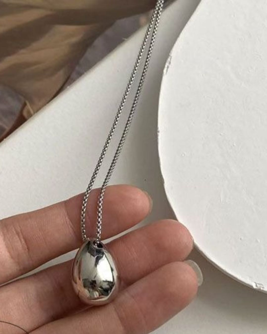 Close-up of fingers holding a highly reflective silver 3D waterdrop pendant necklace, showing its smooth, curved surface.