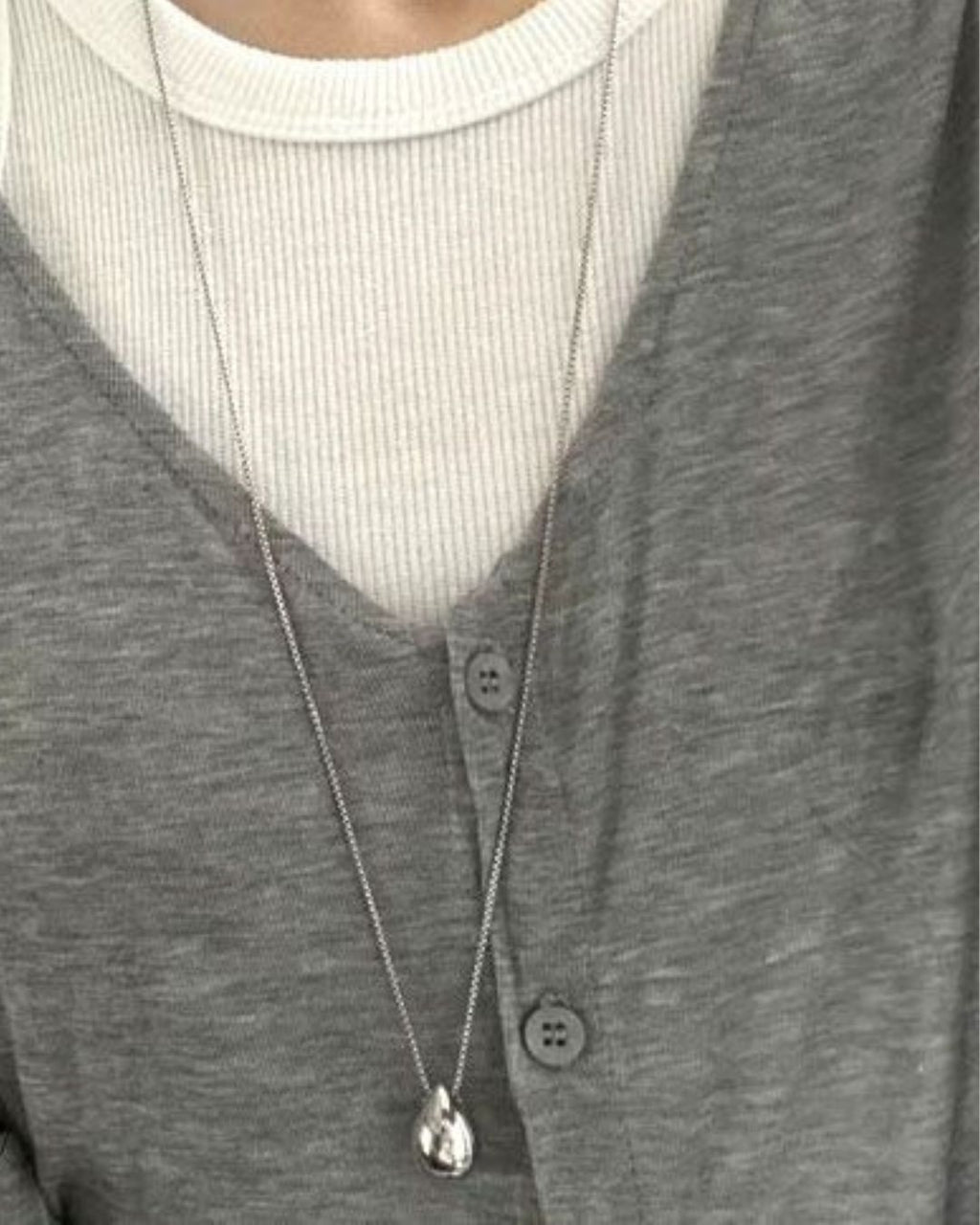 A person wearing a silver teardrop pendant on a long chain layered comfortably over a grey button-up top and white ribbed undershirt.