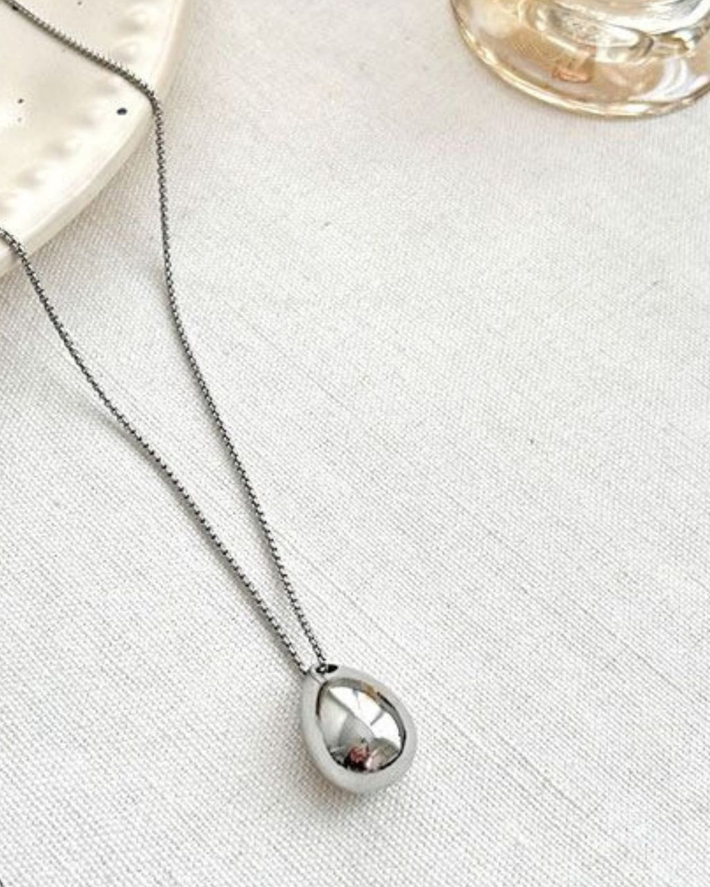 A shiny silver waterdrop pendant necklace resting elegantly on white fabric, with the base of a glass perfume bottle visible in the top right corner.