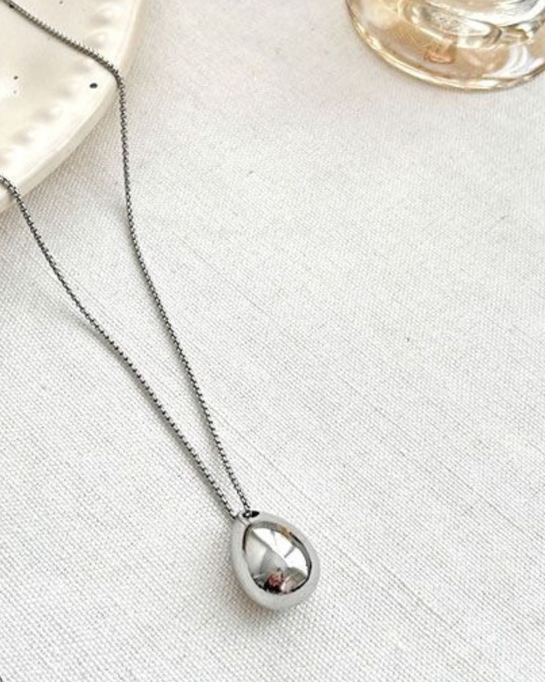 A shiny silver waterdrop pendant necklace resting elegantly on white fabric, with the base of a glass perfume bottle visible in the top right corner.