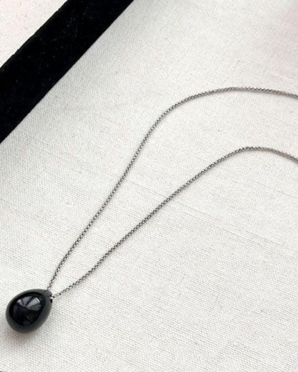 A glossy black teardrop pendant on a delicate silver chain lying flat against a white fabric background near a black velvet edge.