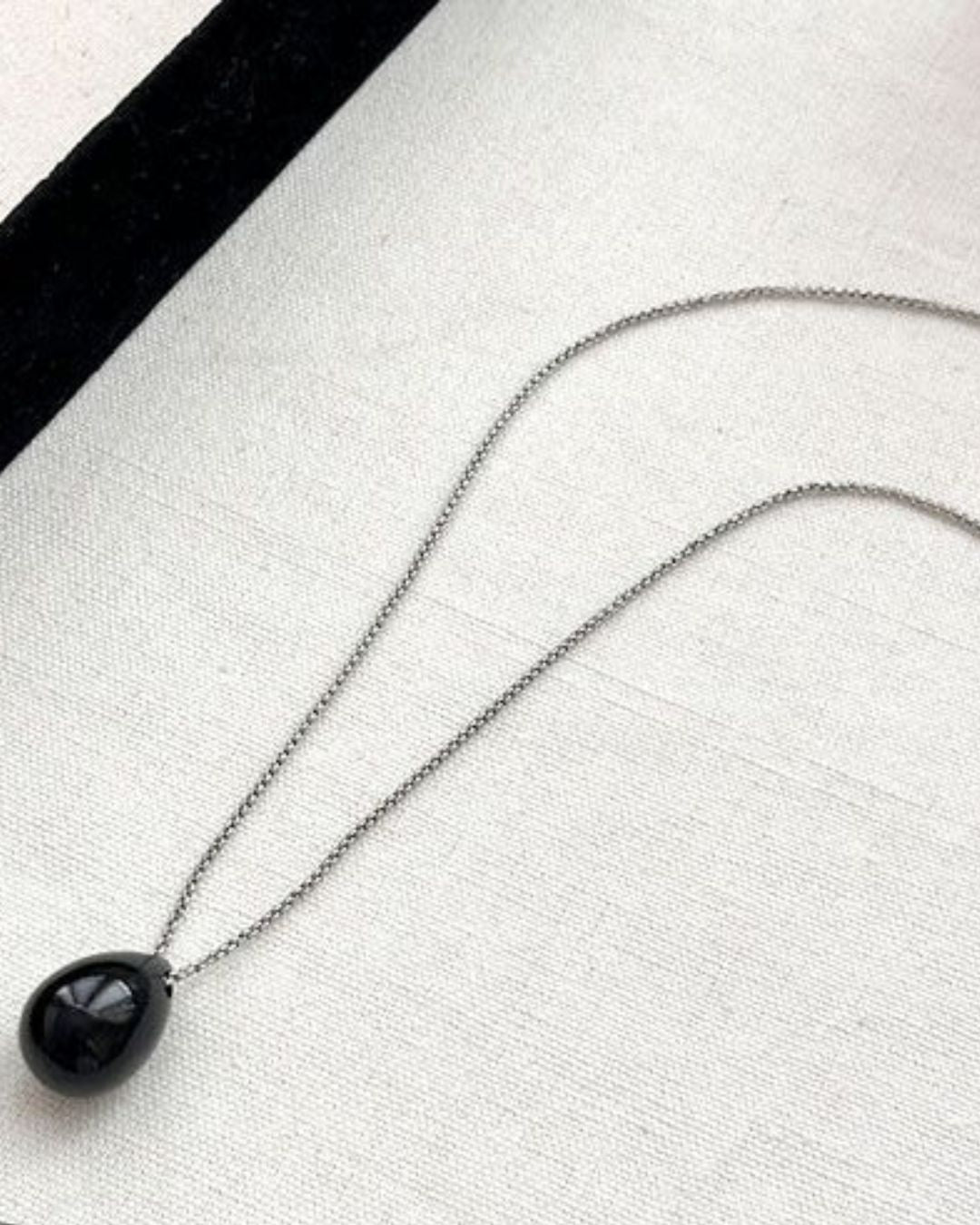 A glossy black teardrop pendant on a delicate silver chain lying flat against a white fabric background near a black velvet edge.