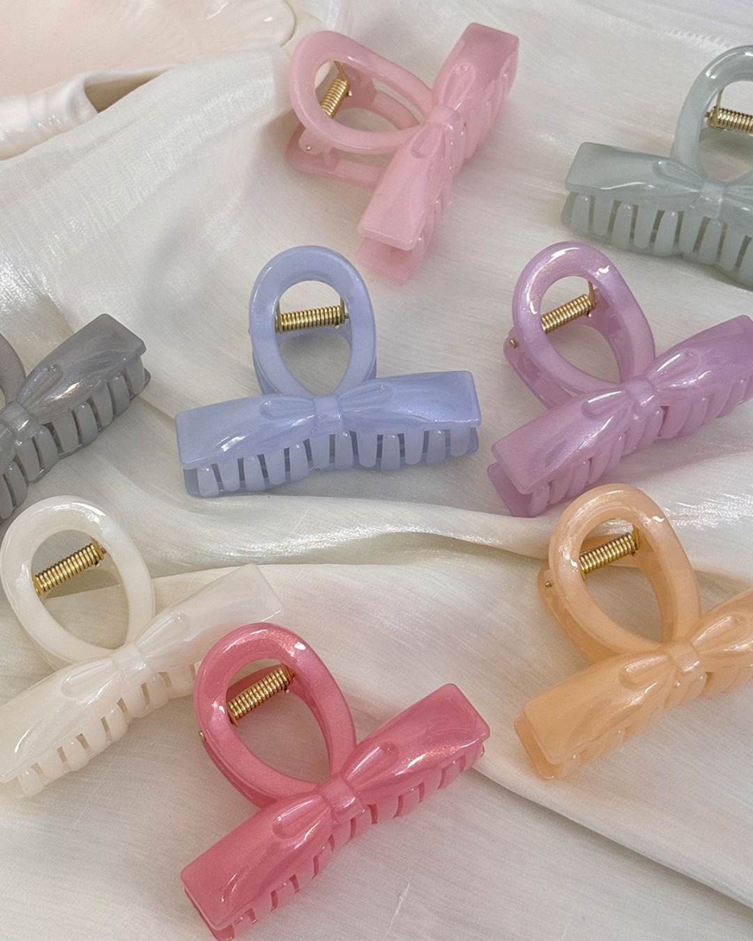 Close-up of four glossy acrylic bow hair clips in slate grey, periwinkle blue, pearl white, and shimmering pink resting on textured white silk fabric.