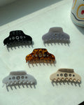 Five trendy hair claw clips with metal eyelet grommets arranged on a white windowsill in direct sunlight. Colors include black, lavender, tortoiseshell, white, and beige.