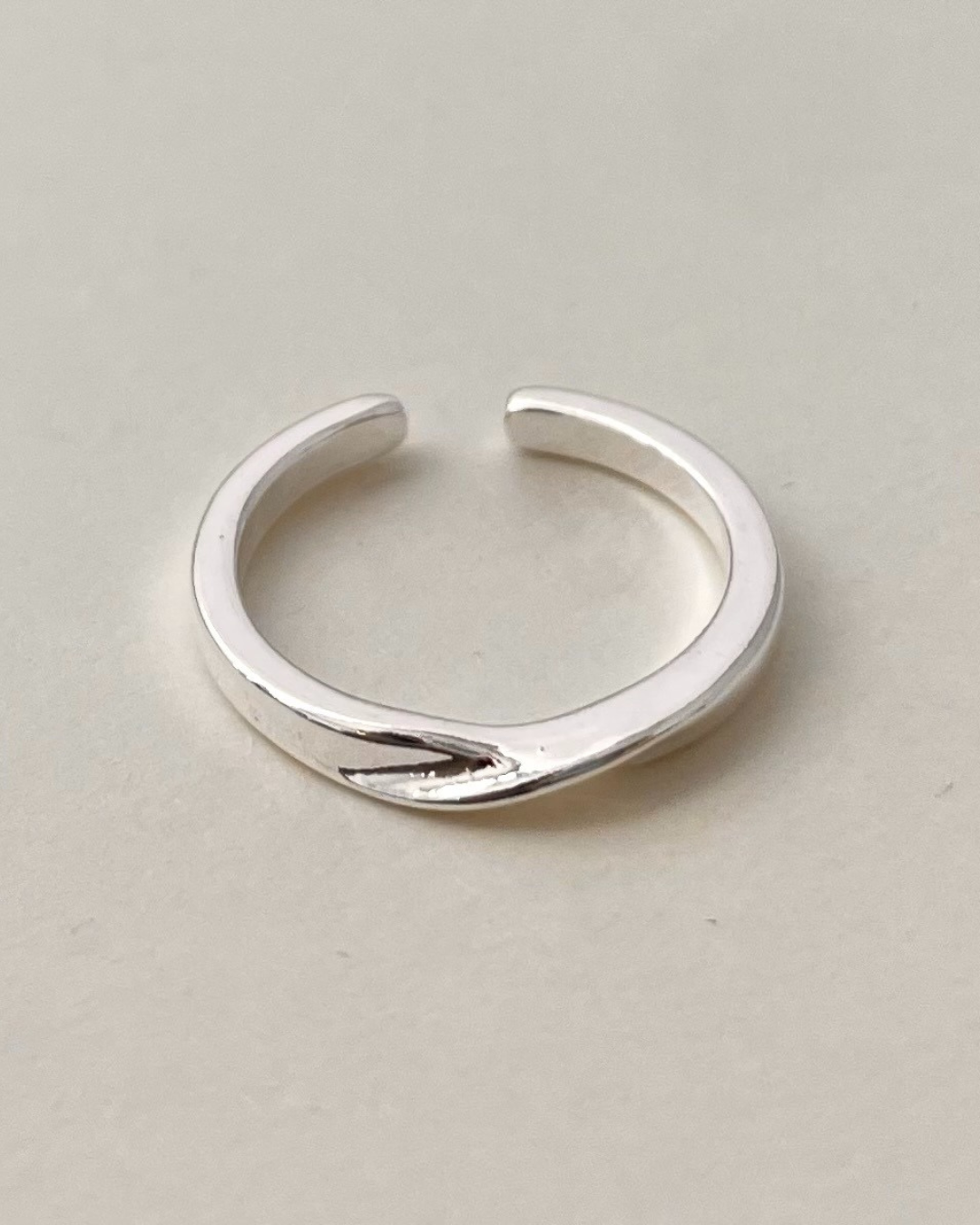 Close-up of a smooth, high-polish silver adjustable open ring featuring a modern sculptural twist detail on a neutral beige background.
