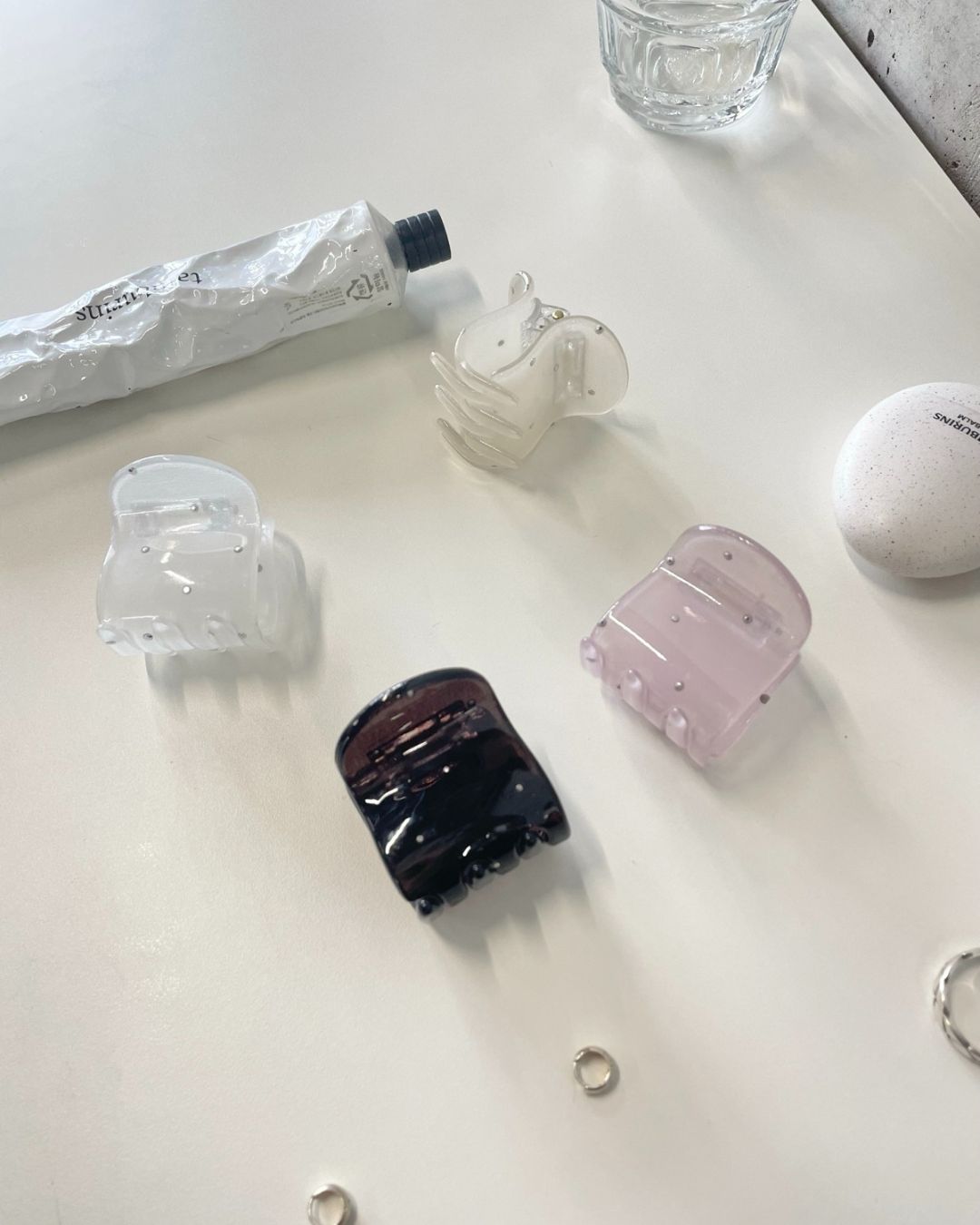 Four square rhinestone hair claw clips in frosted white, milky ivory, translucent pink, and dark brown scattered on a white desk next to a tube of Tamburins hand cream.