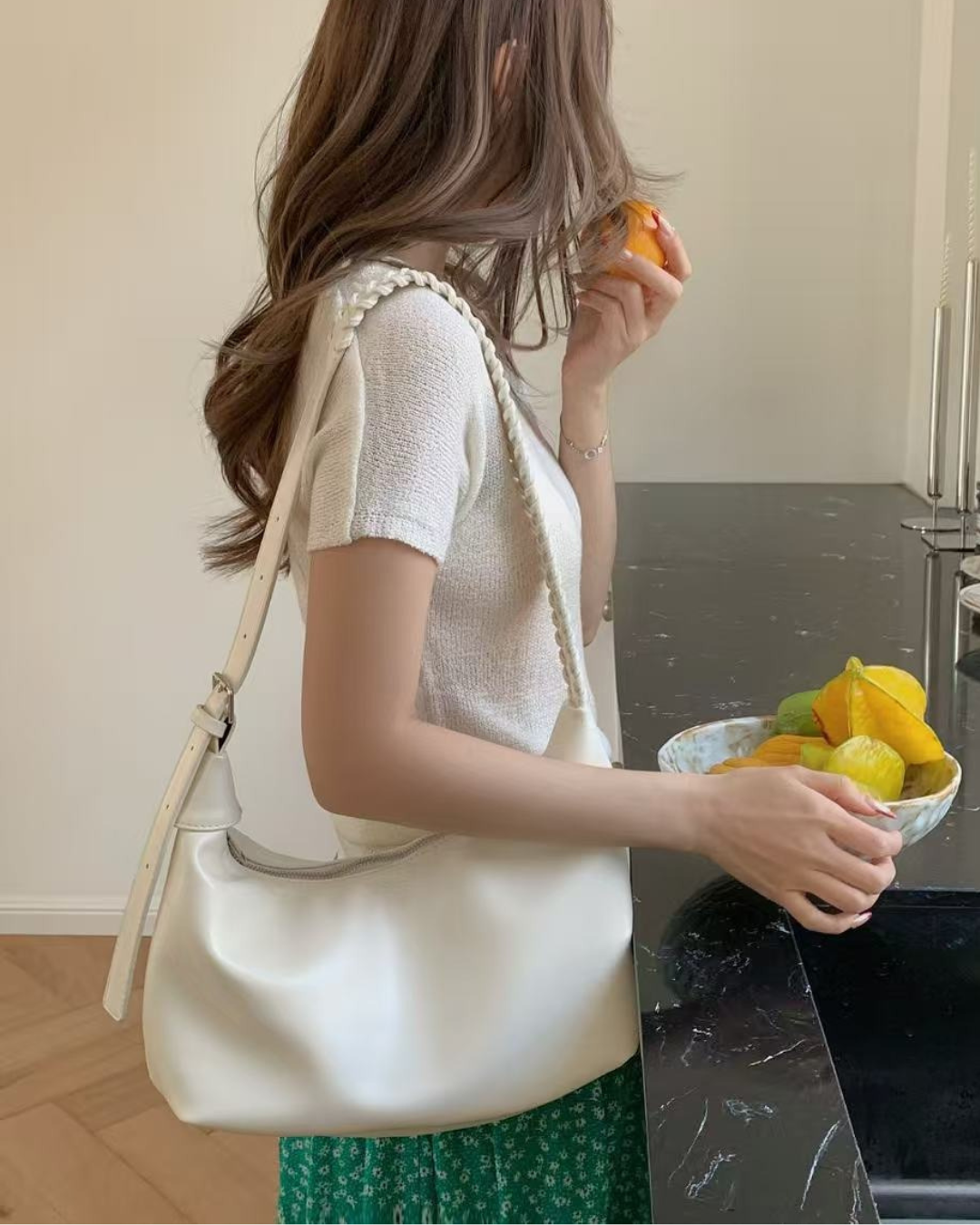 A woman in a modern kitchen holding a bowl of fresh fruit, wearing a cream white minimalist shoulder bag with a subtle braided strap over a white knit top.
