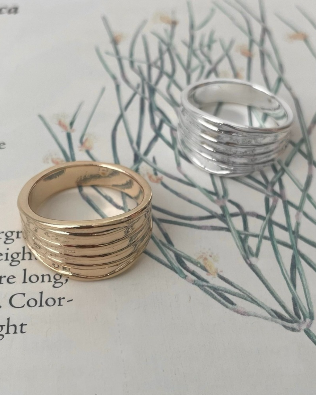 A gold and a silver textured ribbed wide band ring sitting together on an open botanical book featuring green plant illustrations and vintage text.