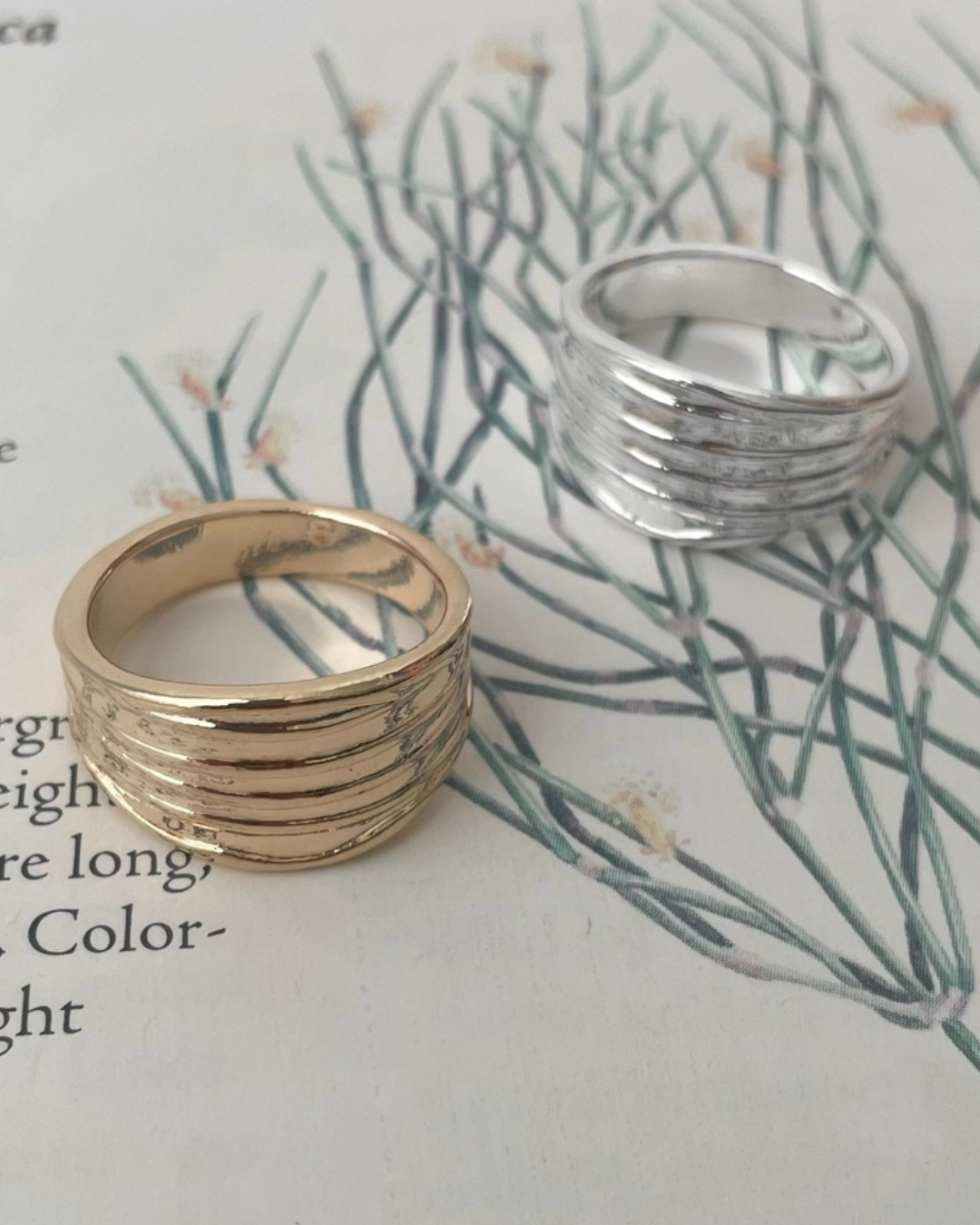 A gold and a silver textured ribbed wide band ring sitting together on an open botanical book featuring green plant illustrations and vintage text.