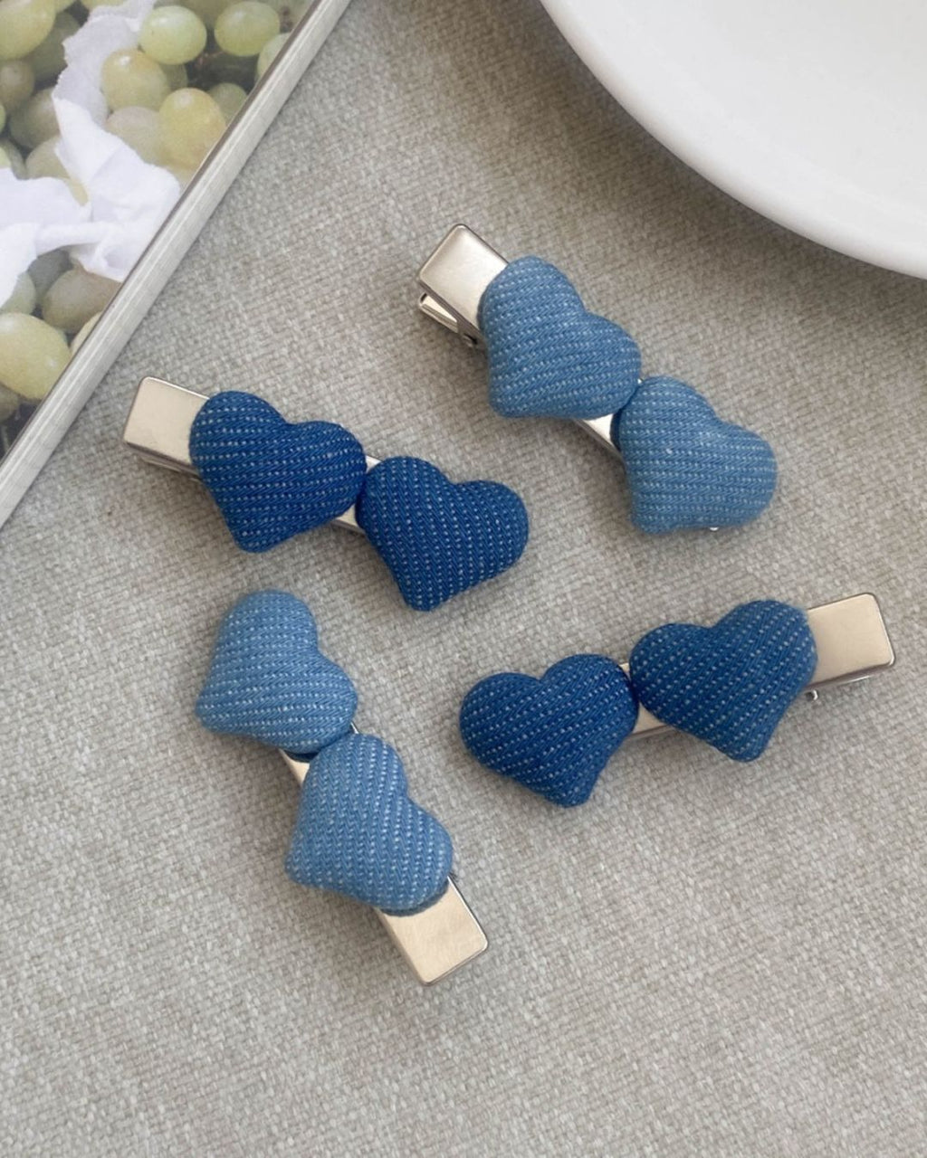 Four puffy denim heart alligator barrettes displayed on a textured beige fabric next to a white plate and a magazine featuring green grapes.