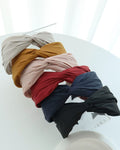 A stack of six wide twisted headbands on a clear acrylic display stand, featuring sand beige, mustard yellow, blush pink, deep red, navy blue, and black.