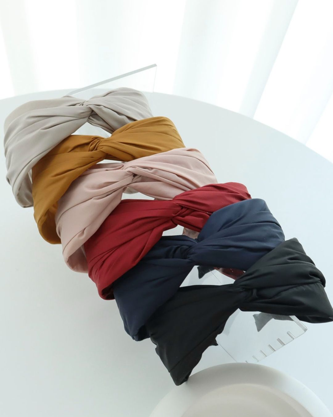 A stack of six wide twisted headbands on a clear acrylic display stand, featuring sand beige, mustard yellow, blush pink, deep red, navy blue, and black.