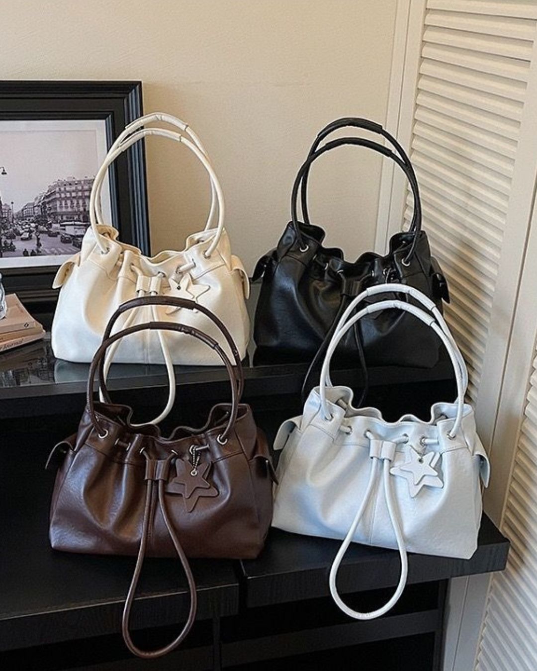 A collection of four faux leather drawstring shoulder bags with star charms, displayed in cream, black, chocolate brown, and light blue on a black shelf.