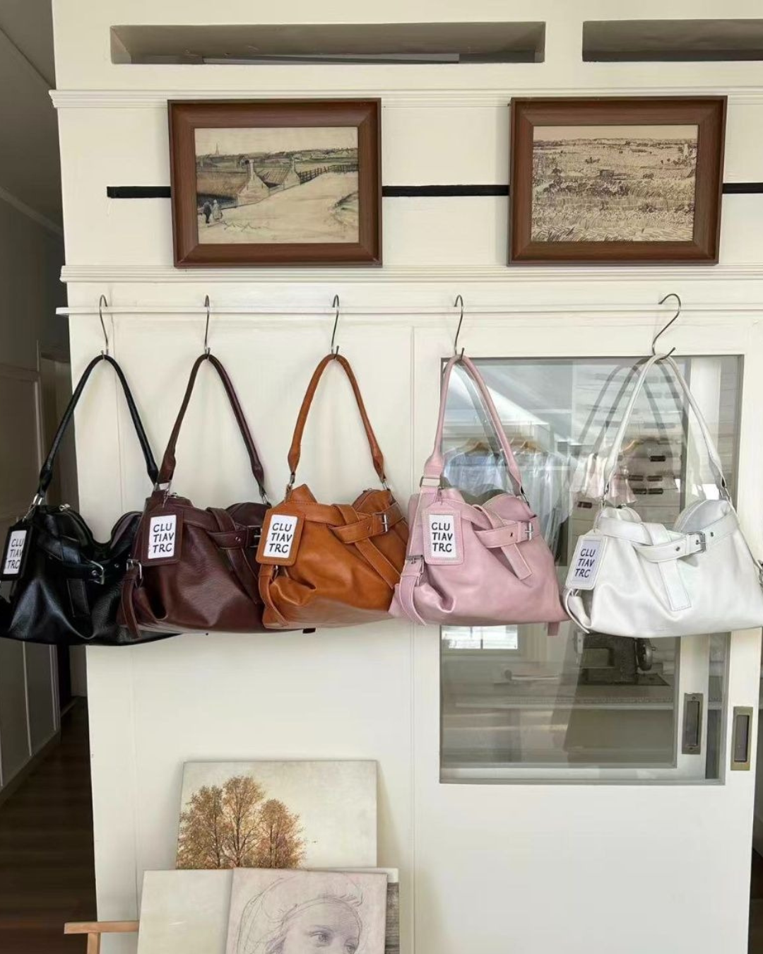 Five soft leather slouchy hobo bags lined up on wall hooks in black, dark espresso brown, caramel tan, blush pink, and cream white.