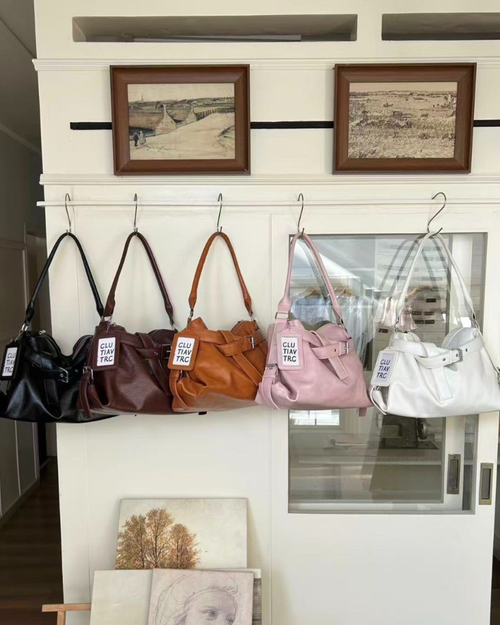 Five soft leather slouchy hobo bags lined up on wall hooks in black, dark espresso brown, caramel tan, blush pink, and cream white.