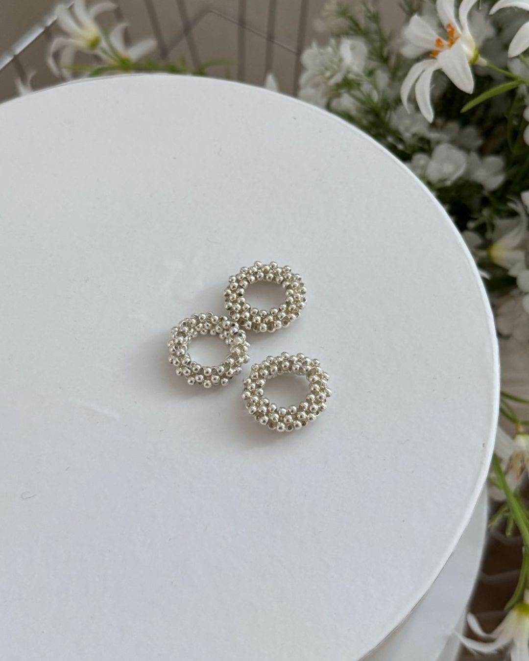 Three chunky silver ball beaded stretch rings clustered together on top of a white round box, with delicate white artificial flowers in the background.