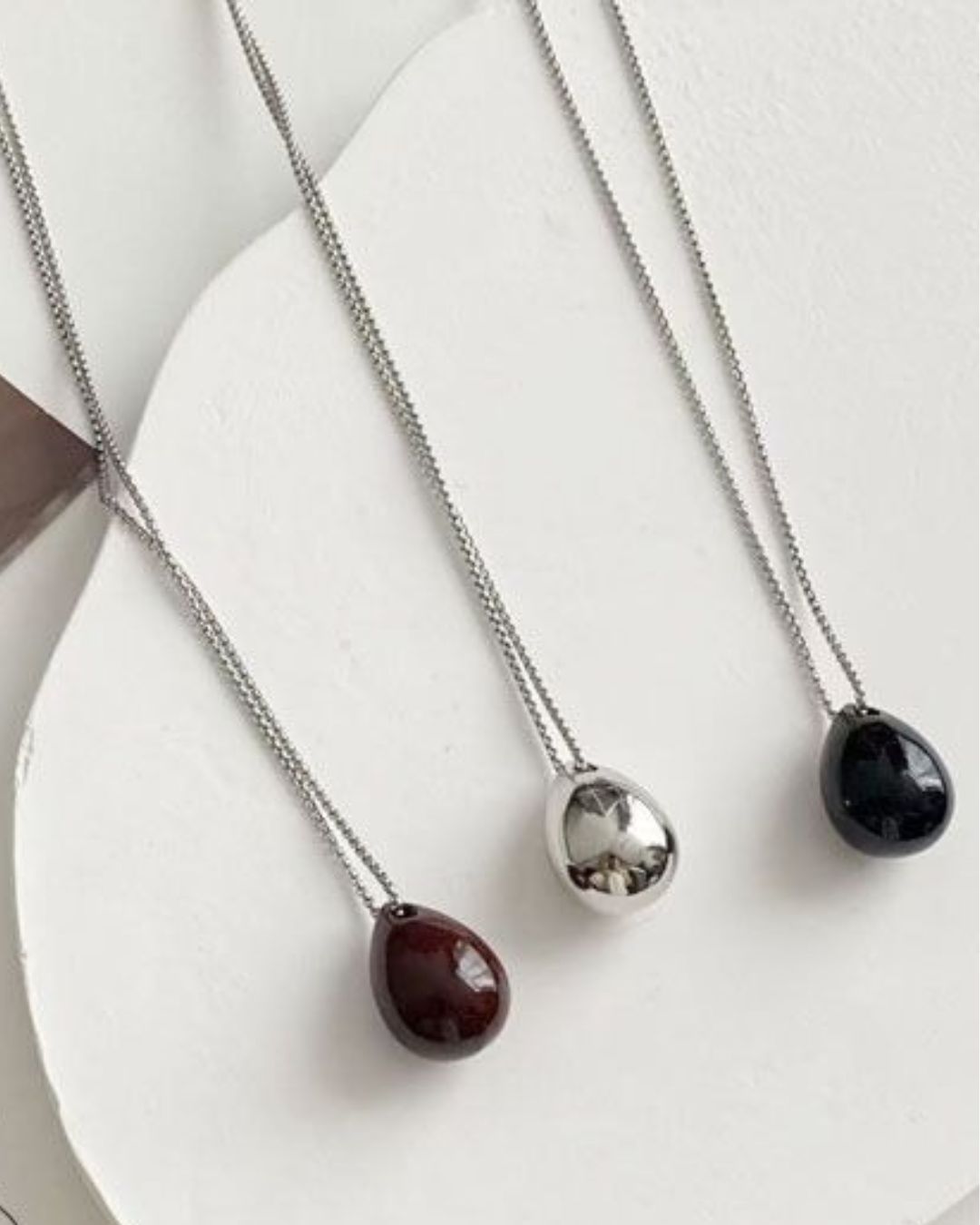 Three sculptural teardrop pendant necklaces in deep burgundy, reflective silver, and glossy black, resting on a textured white display slab.