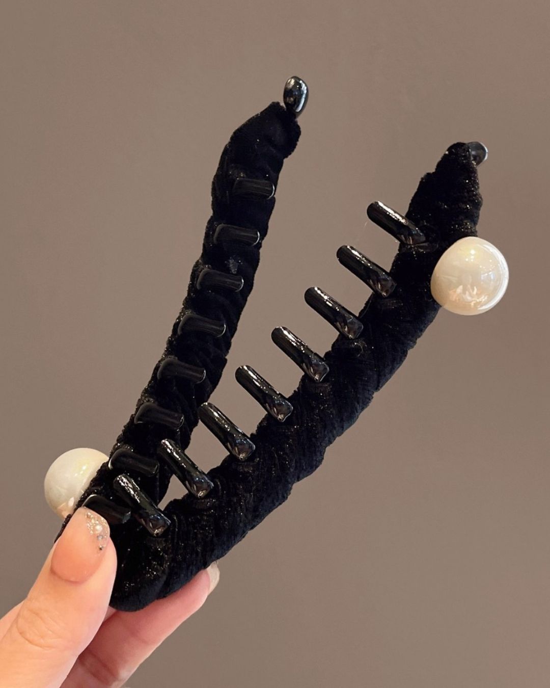 A hand holding an open black velvet banana hair clip, showcasing the large white faux pearl accents and glossy black interlocking inner teeth against a taupe background.