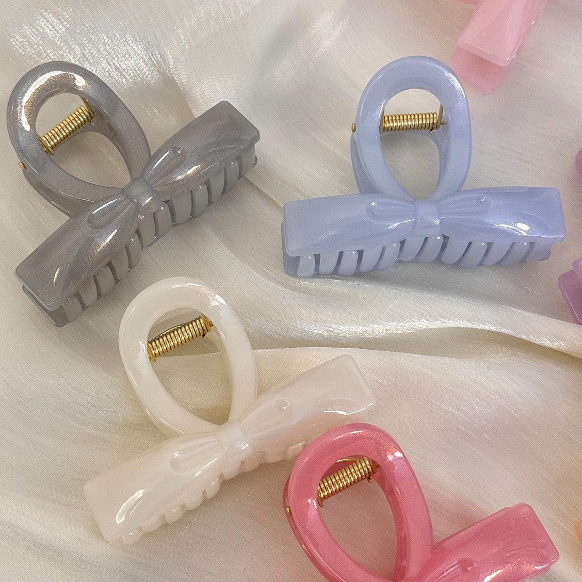 A colorful assortment of seven pearlescent bow claw clips in pink, peach, lilac, periwinkle, slate grey, white, and mint scattered on draped white silk.