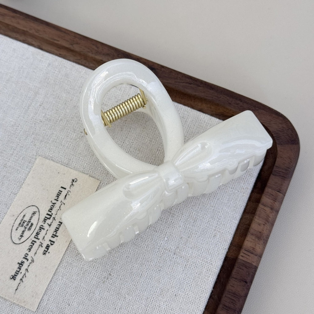 A pearl white shimmering flat ribbon hair claw clip sitting elegantly on a vintage text fabric tag and wooden tray.