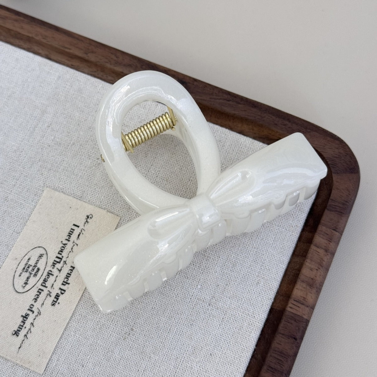 A pearl white shimmering flat ribbon hair claw clip sitting elegantly on a vintage text fabric tag and wooden tray.