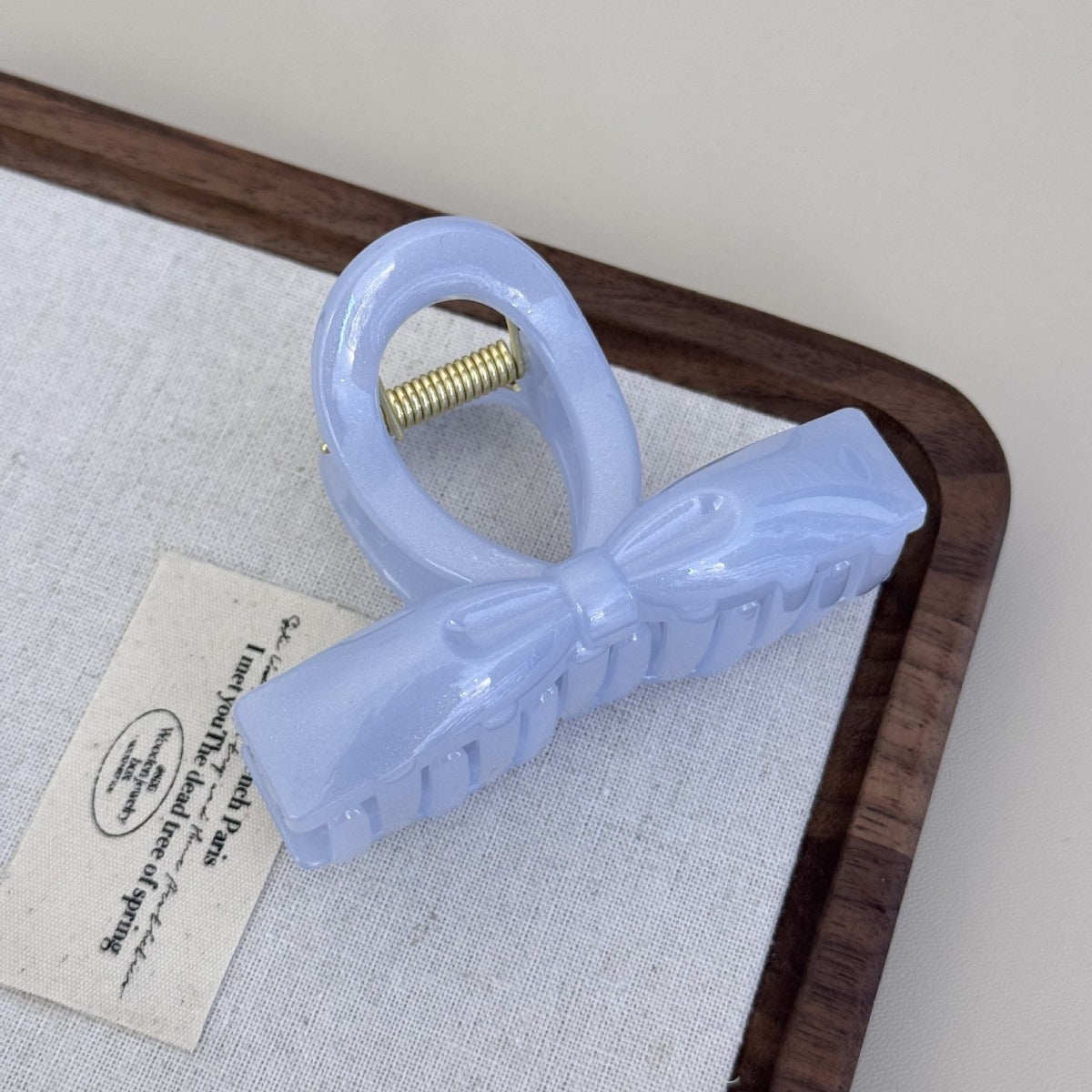 A periwinkle blue glossy acrylic bow claw clip displayed on a white textured fabric background with a wooden frame.