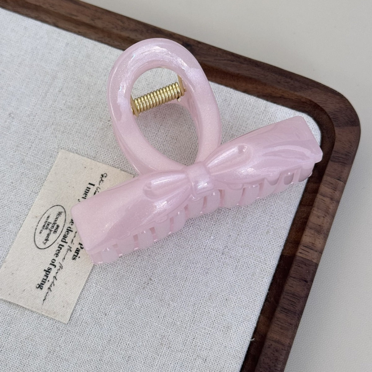 Top-down view of a light pink structured ribbon hair claw clip featuring a luminous pearlescent finish on a white fabric mat.
