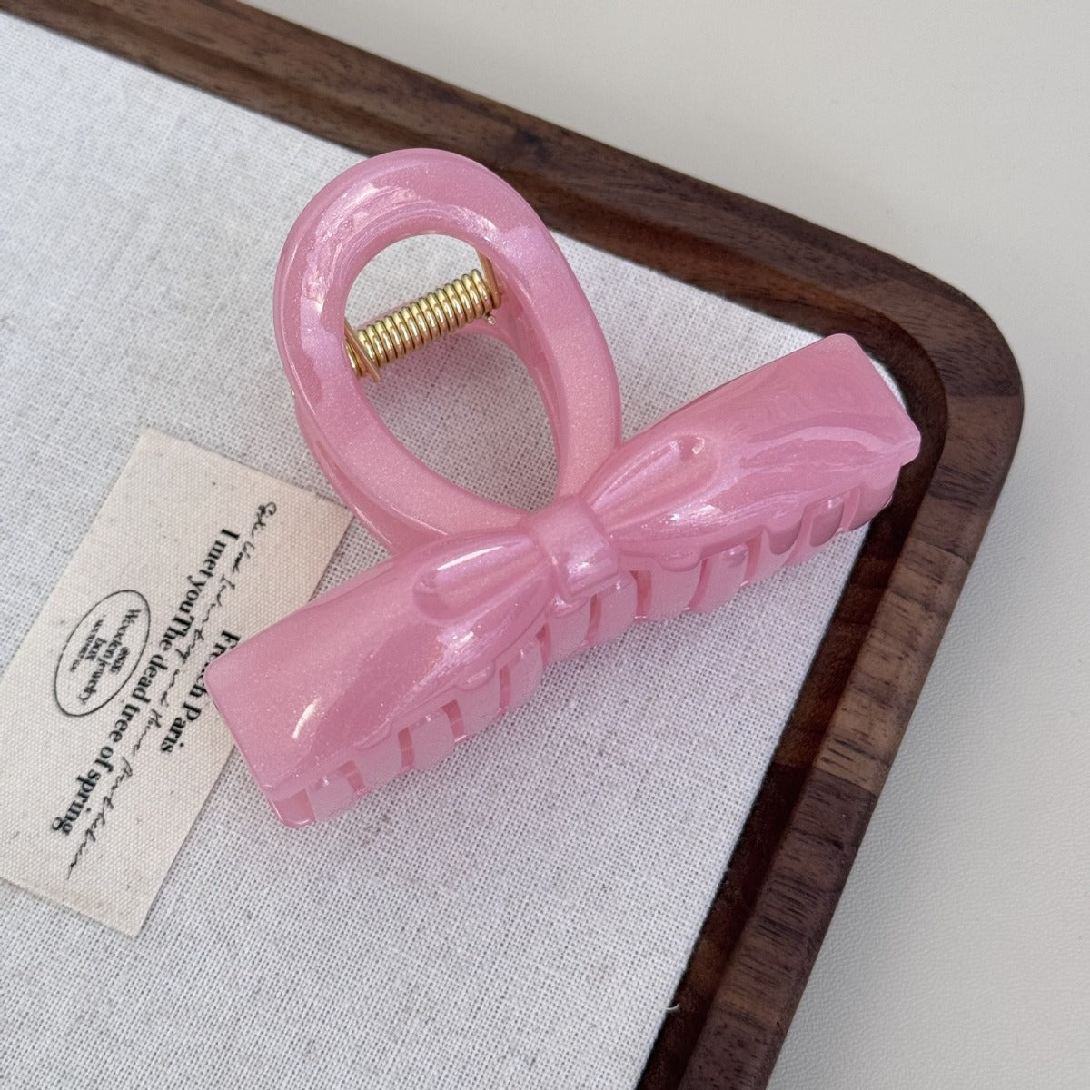 A shimmering pink pearlescent flat ribbon bow claw clip resting on a rustic white fabric and wood display tray.