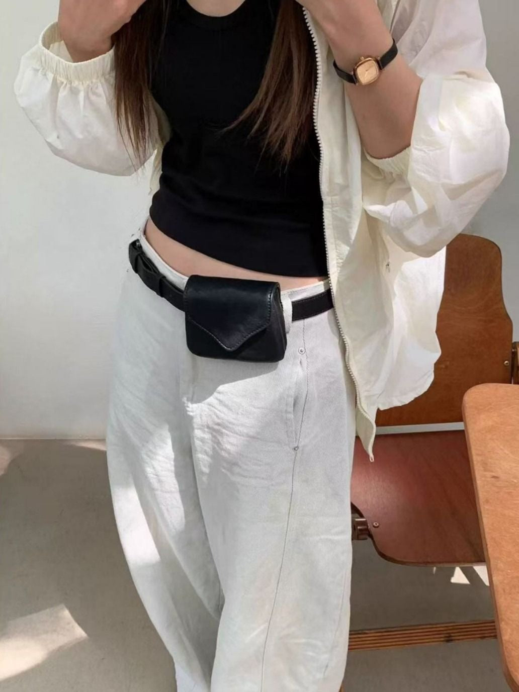 Person wearing a white jacket, black top, and light-colored pants with a black waist bag indoors.