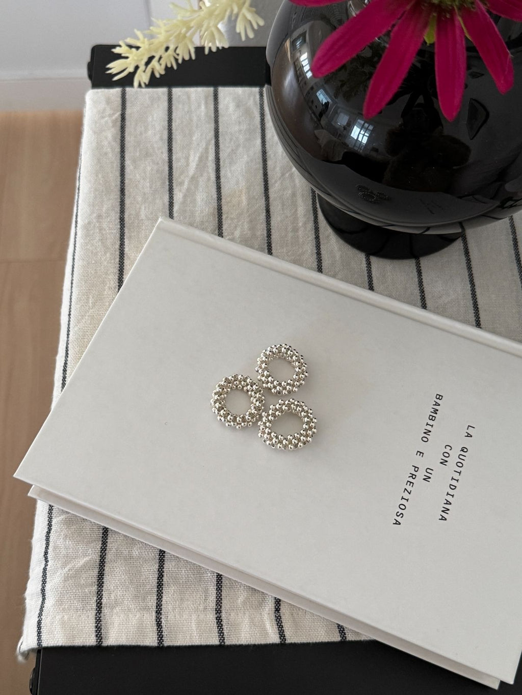 Three silver textured ball rings resting on the cover of a white book titled "La Quotidiana" placed on a black and white striped tablecloth.