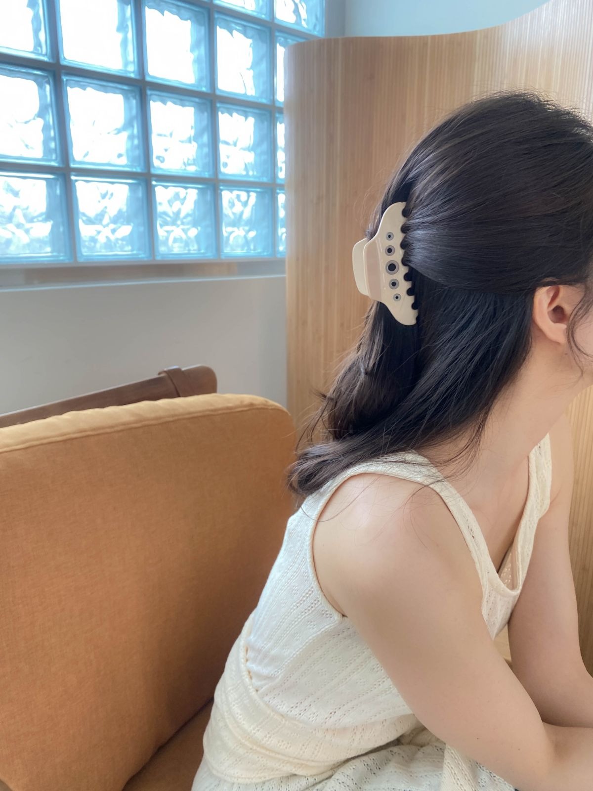 A woman with dark hair styled in a half-updo wearing a beige acrylic hair claw clip with silver eyelet details, sitting in a bright room with glass block windows.