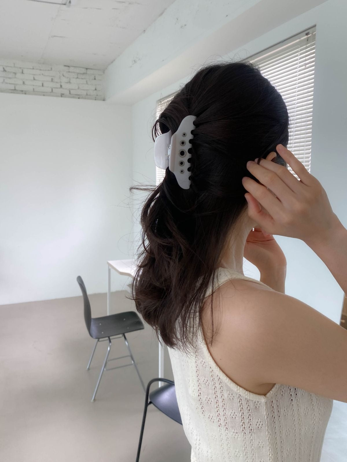 A woman adjusting her dark wavy hair, secured in a messy half-updo with a white eyelet detail hair claw clip, standing in a minimalist white room.