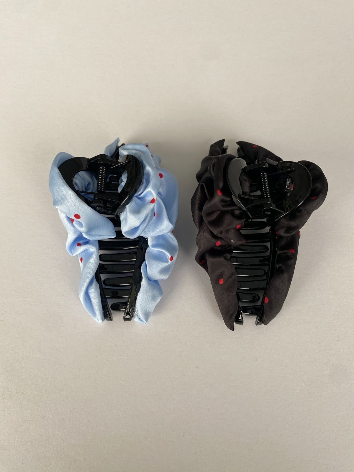 Top-down view showing the inner plastic teeth of two satin scrunchie hair claw clips, one light blue and one black, both featuring small red polka dots.