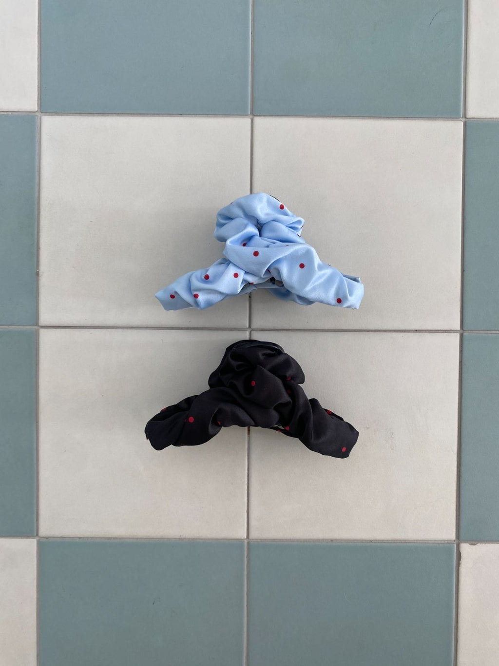 A light blue and a black ruched fabric hair claw clip with red polka dots resting flat on a light blue and white checkered tile background.