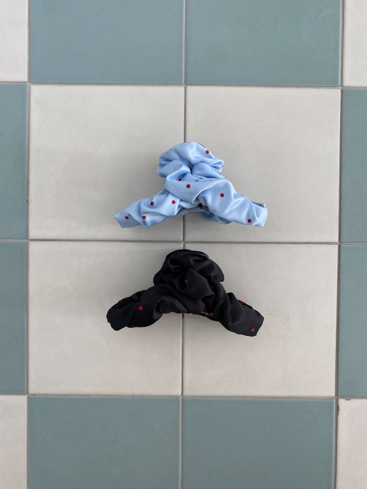 A light blue and a black ruched fabric hair claw clip with red polka dots resting flat on a light blue and white checkered tile background.