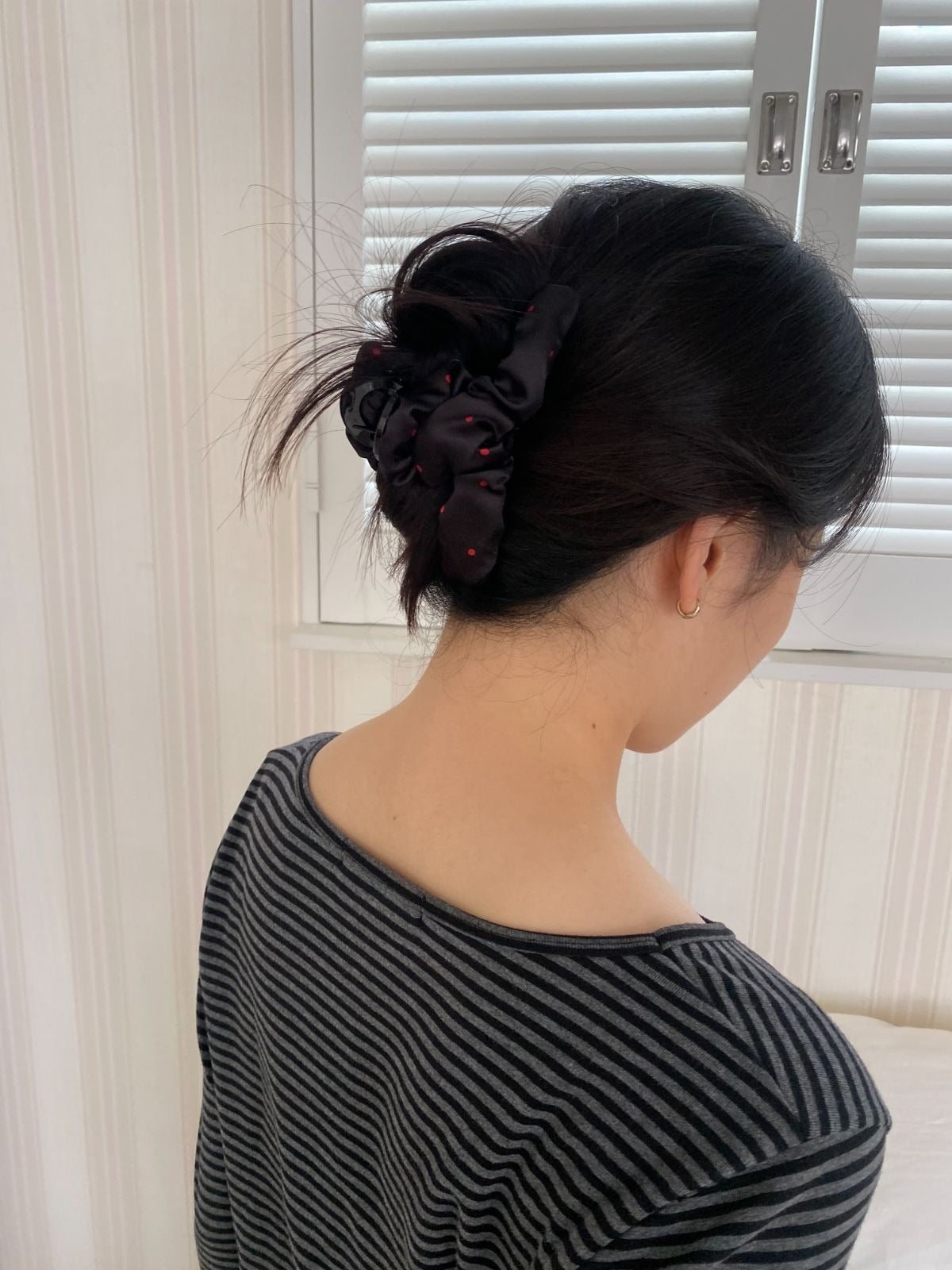 Back view of a woman wearing a black and red polka dot scrunchie claw clip holding her dark hair in a messy updo, wearing a grey striped top.