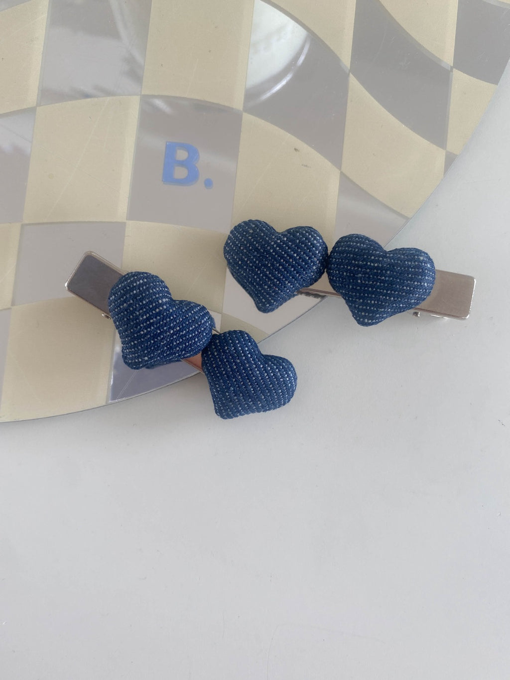 Two dark blue puffy denim heart hair clips resting on a trendy tan and white checkered mirror tray.