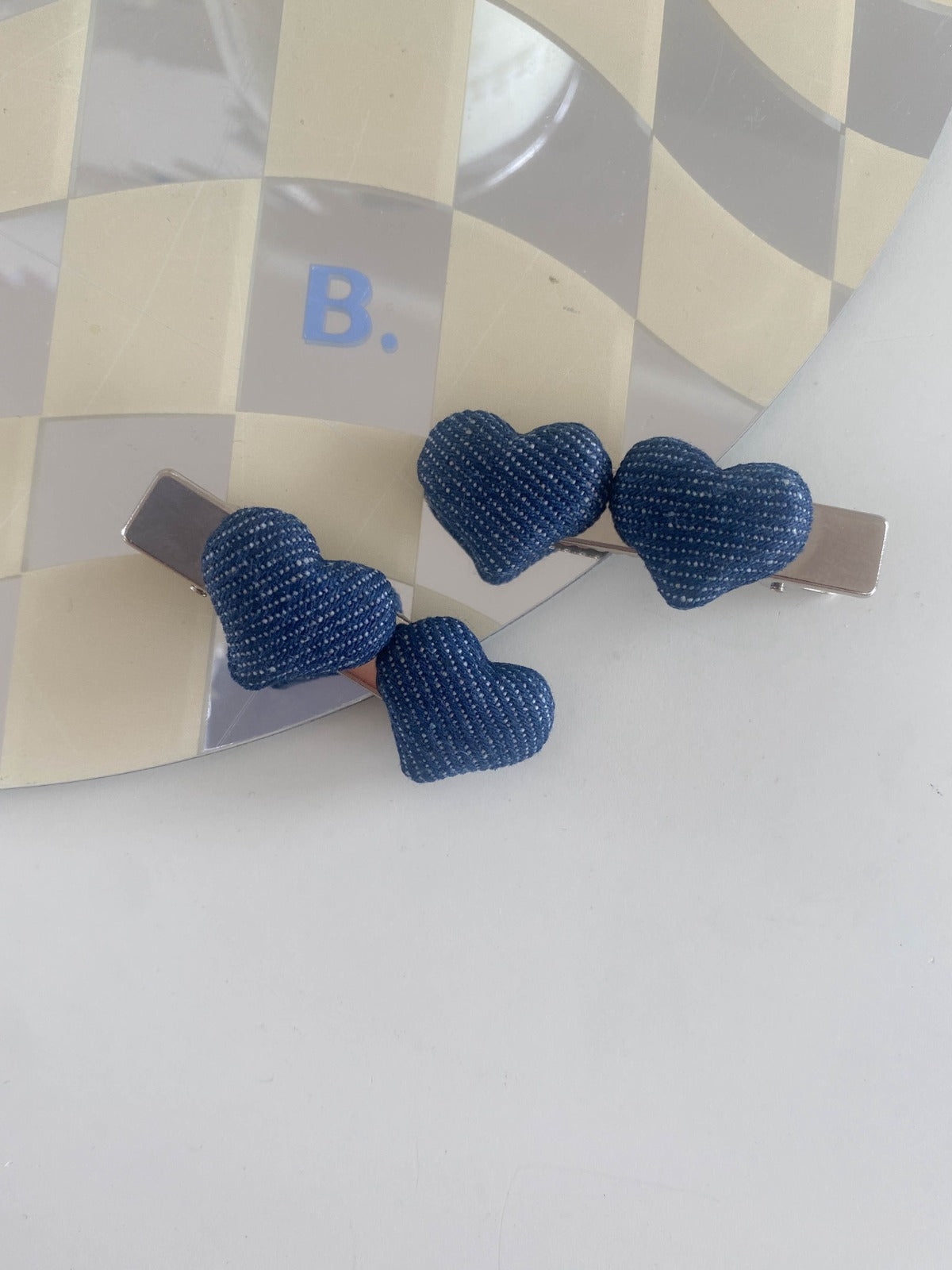 Two dark blue puffy denim heart hair clips resting on a trendy tan and white checkered mirror tray.