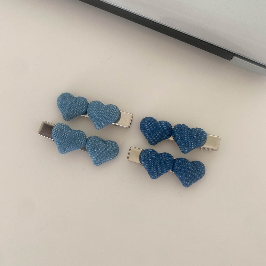 Four twin-heart denim hair clips—two in a light wash and two in a dark wash—sitting on a minimalist white desk next to a laptop.