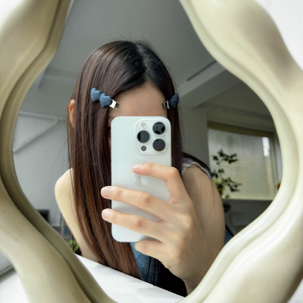 A woman taking a mirror selfie, using two dark denim heart hair clips to stylishly pin back her front layers.