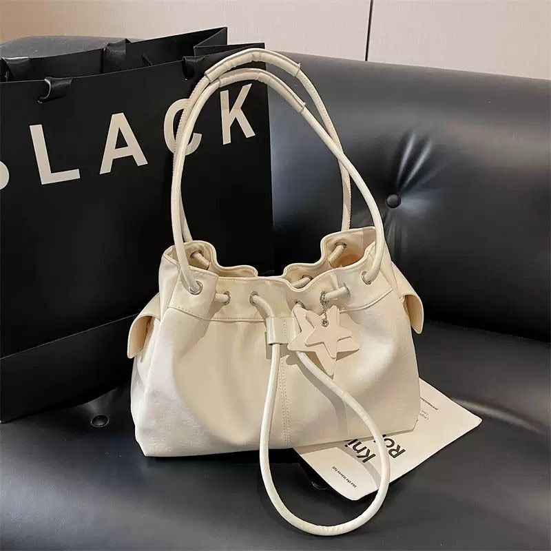 Cream white slouchy faux leather shoulder bag featuring a ruched drawstring top and a matching star charm, resting on a black tufted leather sofa.