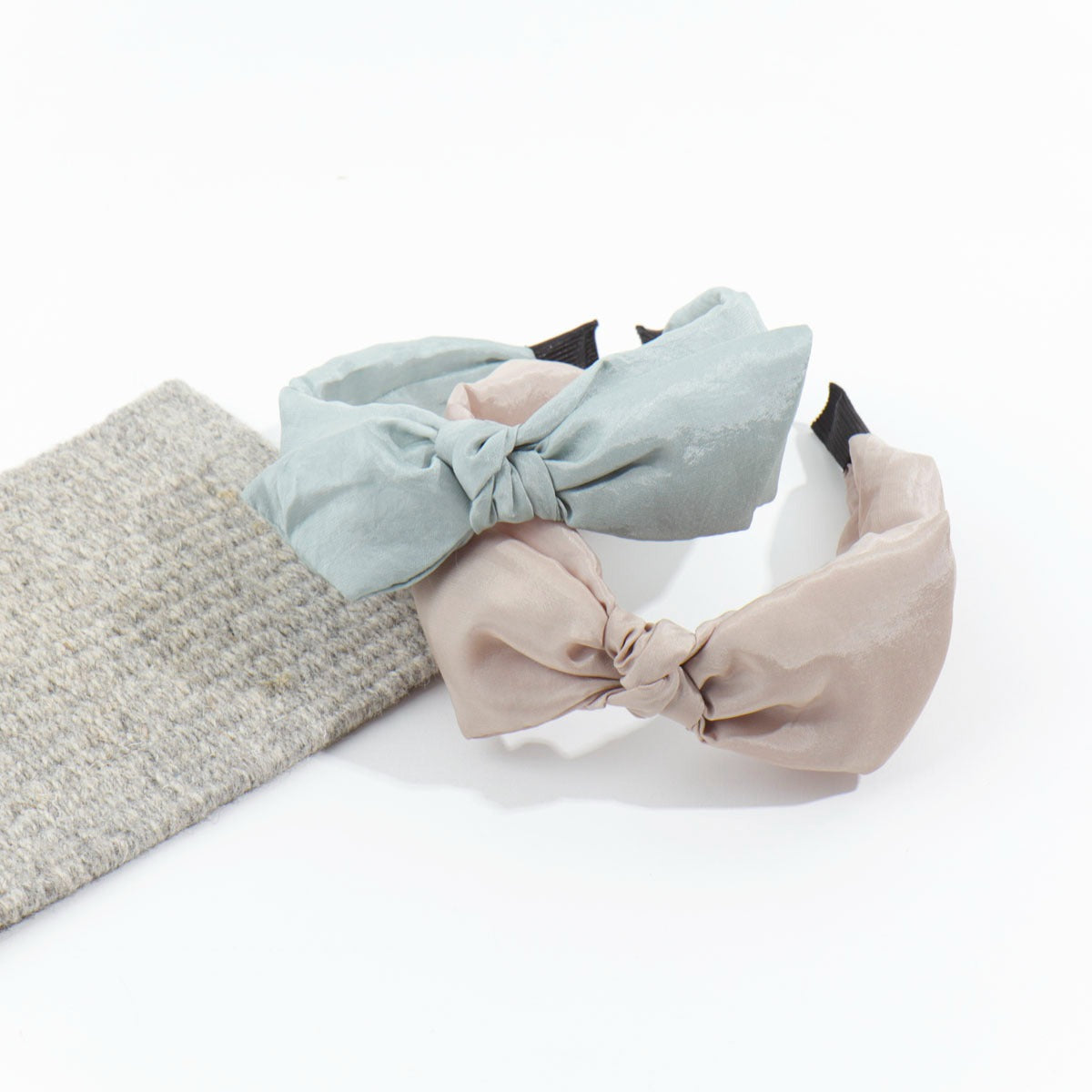 Two silk-feel knotted headbands in sage green and warm beige resting gently next to a textured grey fabric swatch.