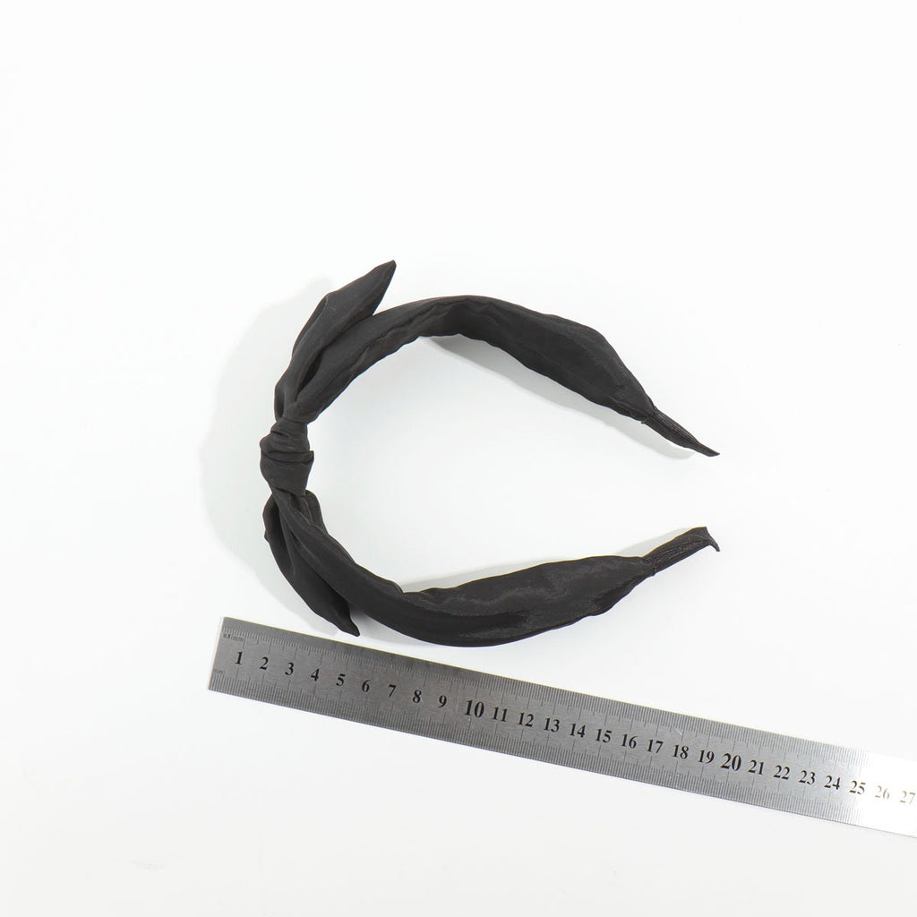 Top-down view of a black satin bow headband placed next to a silver metal measuring ruler to display its standard sizing scale.
