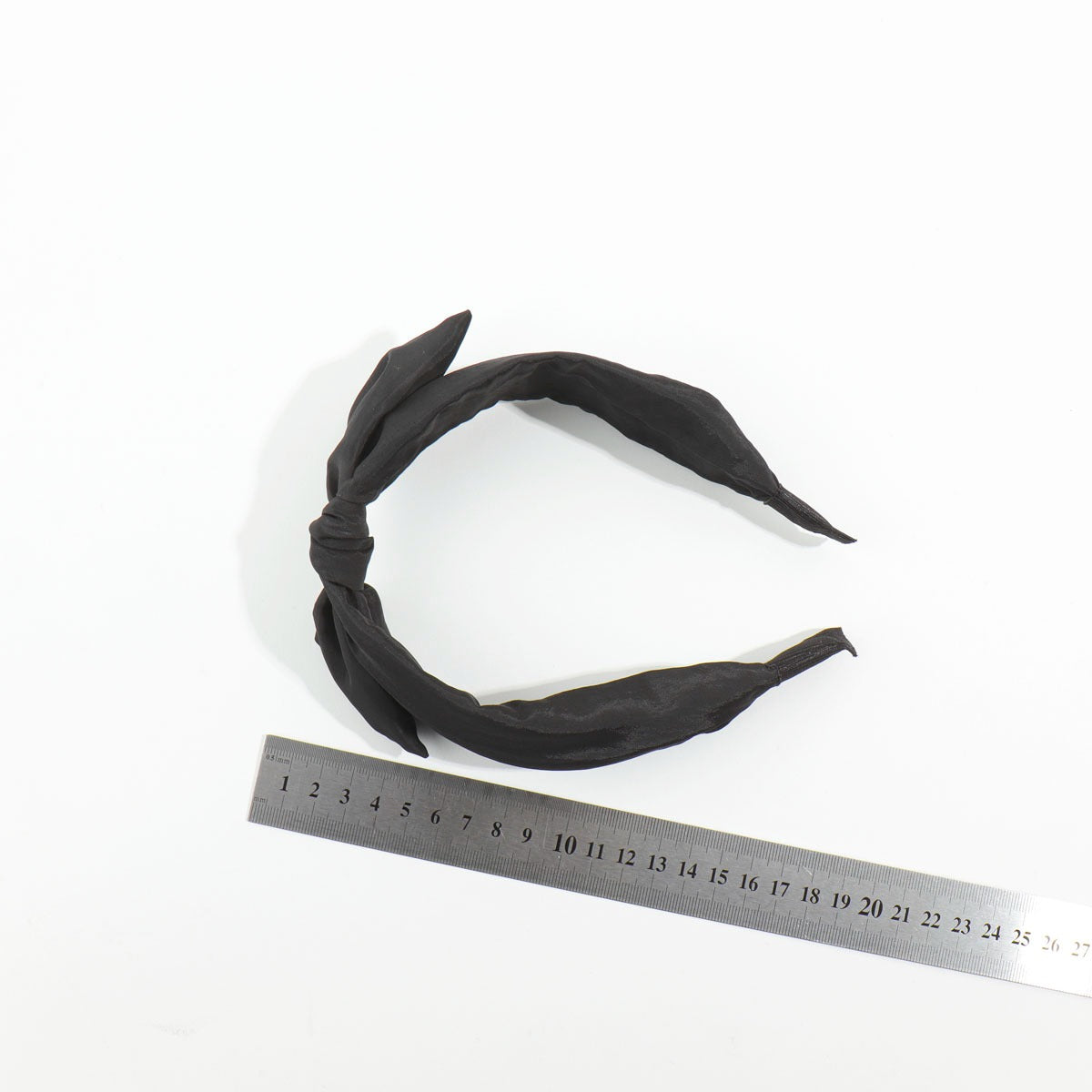 Top-down view of a black satin bow headband placed next to a silver metal measuring ruler to display its standard sizing scale.