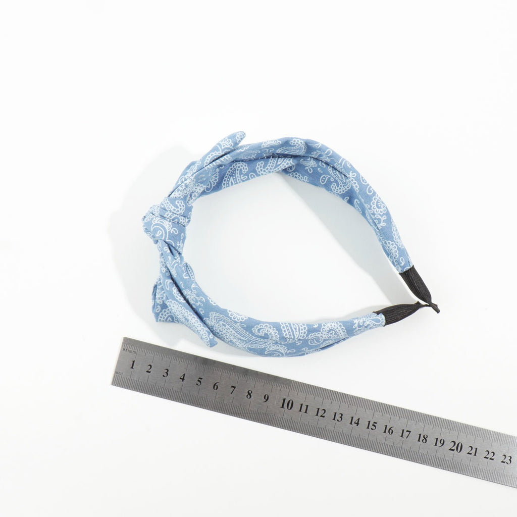 Top-down view of a light blue paisley print knotted headband placed next to a metal measuring ruler to demonstrate sizing.
