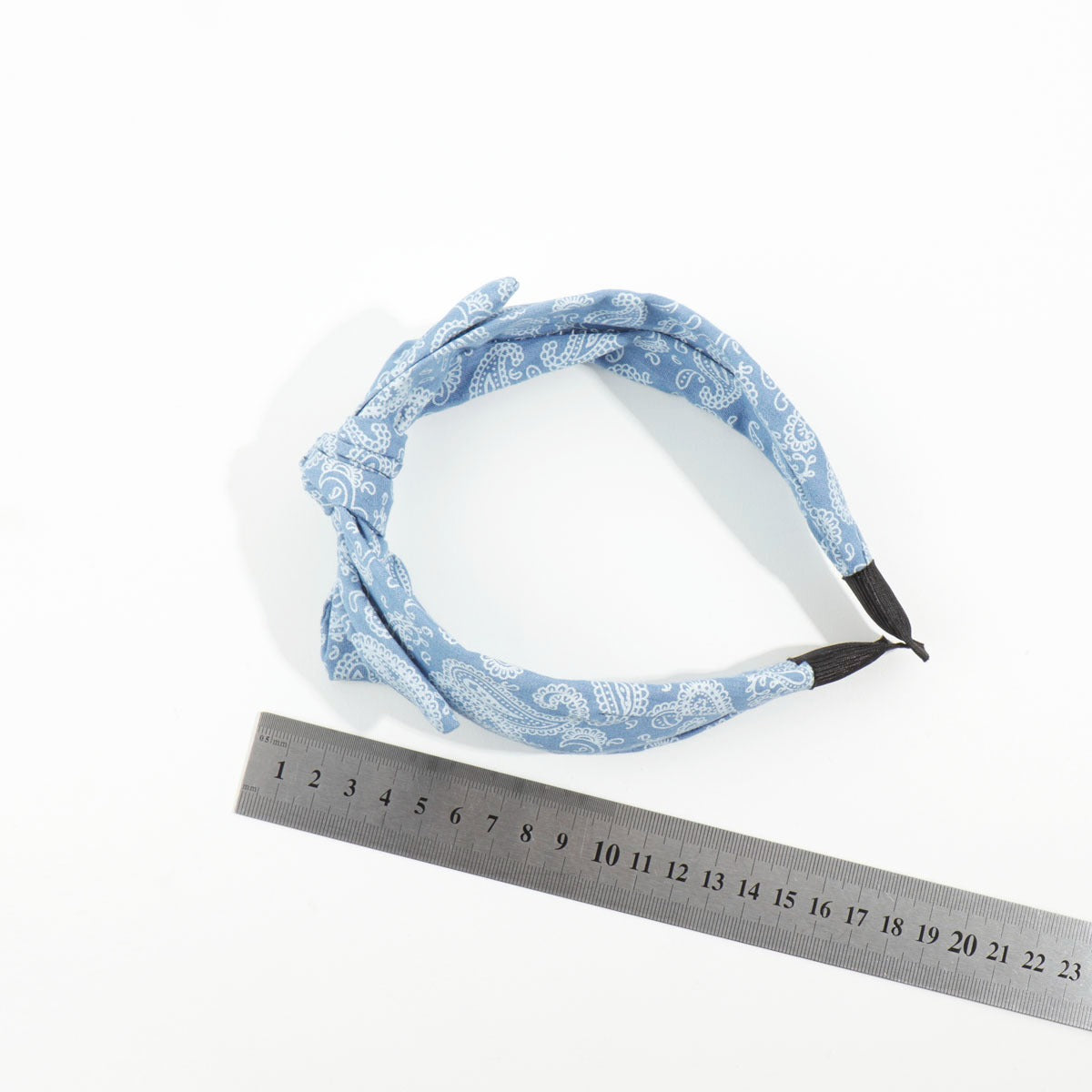 Top-down view of a light blue paisley print knotted headband placed next to a metal measuring ruler to demonstrate sizing.