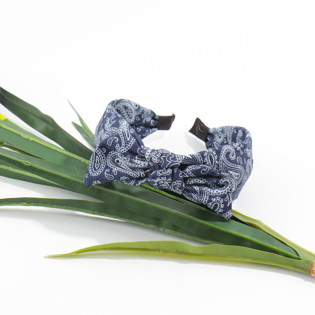 A dark blue bandana print knotted bow headband resting stylishly on top of a long green faux plant stem against a white background.