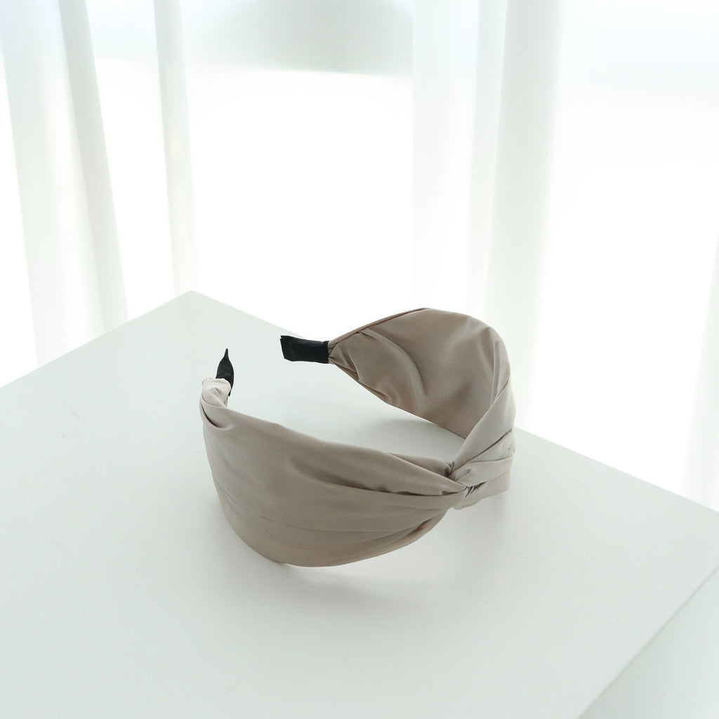 A single sand beige soft matte twisted headband resting gracefully on a white display pedestal with sheer white curtains in the background.