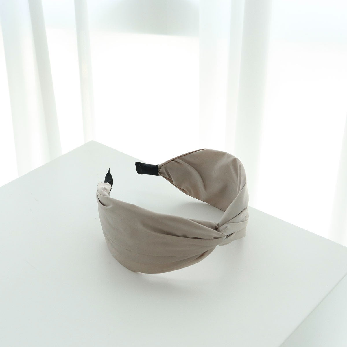 A single sand beige soft matte twisted headband resting gracefully on a white display pedestal with sheer white curtains in the background.